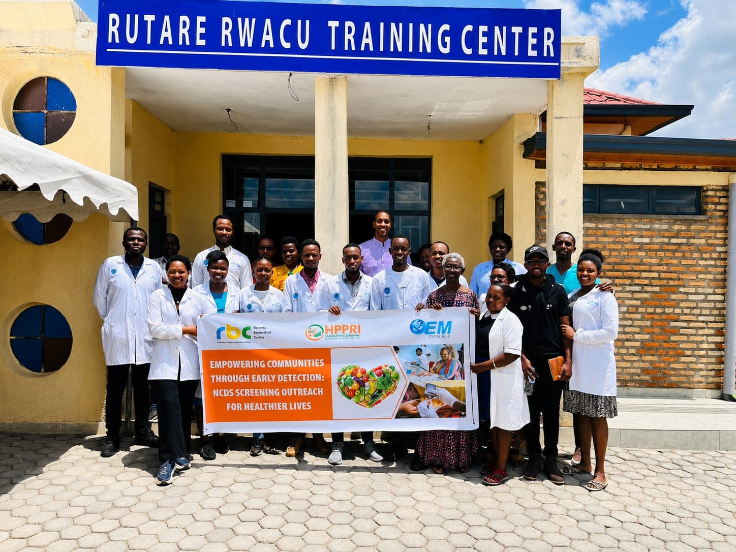 A team in white coats and everyday clothes standing under the Rutare Rwacu Training Center sign, holding a banner that reads “Empowering communities through early detection: NCDs screening outreach for healthier lives”.