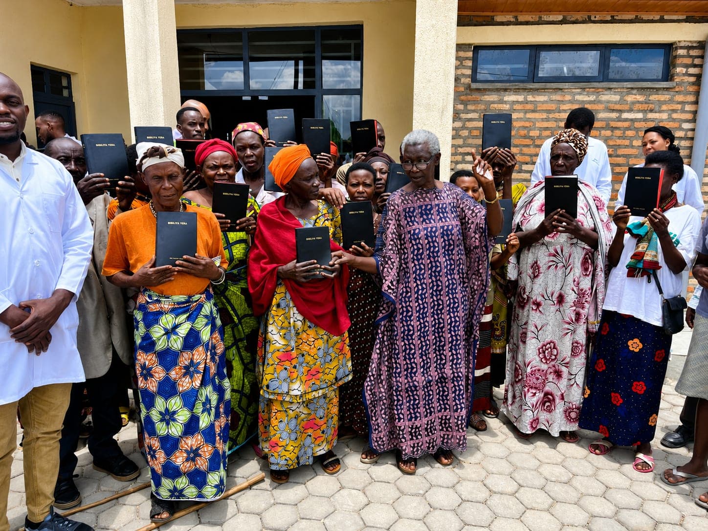 A large group standing outside a brick building, each person holding up a black Kinyarwanda Bible marked Bibiliya Yera.