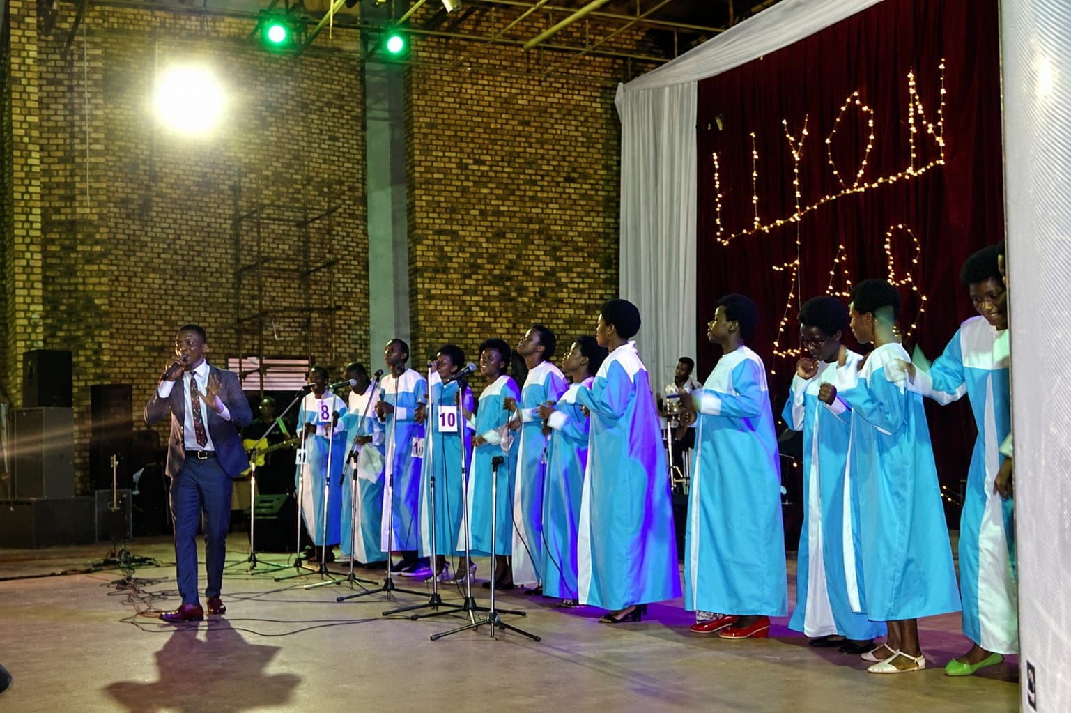 A choir in blue and white robes singing into a row of microphones on a lit stage, a host with a microphone beside them, and the words ELYON XMAS picked out in lights on the backdrop.